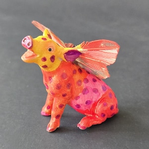 May include: A whimsical, hand-painted figurine of a pig with feathered wings. The pig is primarily orange and pink with purple polka dots. The wings are a mix of red and orange. The pig is sitting with its mouth open.