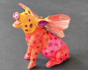 Könnte beinhalten: Eine handbemalte Figur eines Schweins mit gefiederten Flügeln. Das Schwein ist hauptsächlich orange und rosa mit lila Punkten. Die Flügel sind rot und orange. Das Schwein sitzt mit offenem Mund, ein einzigartiges Dekorationsstück.