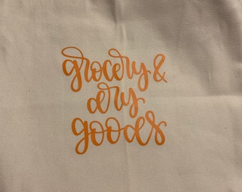 Könnte beinhalten: Eine weiße Stofftasche mit orangefarbenem Text, der "grocery & dry goods" lautet.