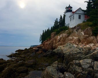 May include: A white lighthouse with a red roof sits on a rocky cliff overlooking the ocean. The sky is cloudy with a bright sun shining through the clouds.