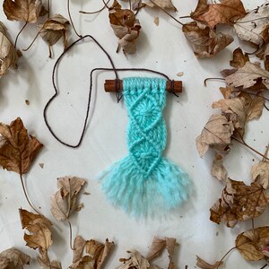 Puede incluir: Un pequeño tapiz de macramé de color azul claro con flecos. El tapiz está sujeto a un palo de canela con cuerda marrón.
