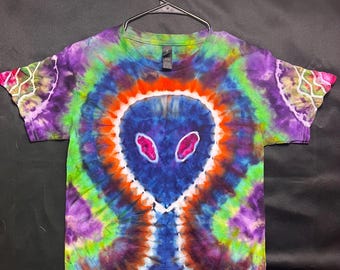Puede incluir: Una camiseta teñida con un diseño alienígena. La camiseta tiene una cara alienígena azul con ojos rosas, rodeada de círculos concéntricos de color naranja, verde, morado y azul. Las mangas tienen un diseño blanco en los bordes.
