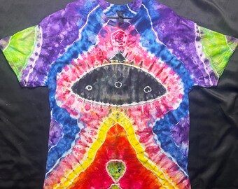Puede incluir: Una camiseta teñida con un diseño psicodélico vibrante. La camiseta presenta una imagen central de un OVNI negro con tres ventanas circulares, rodeada de una colorida gama de tonos rojos, amarillos, azules, morados y verdes. La camiseta está colgada en una percha negra.