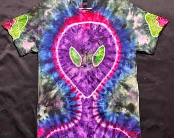 Puede incluir: Una camiseta teñida con un diseño de alienígena morado, ojos verdes y un contorno rosa. La camiseta tiene cuello redondo y mangas cortas. El fondo es una mezcla de colores azul, verde y rosa.