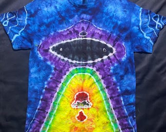 Puede incluir: Una camiseta tie-dye con un diseño de arcoíris y una forma de OVNI en blanco y negro en la parte superior. La camiseta es de color azul oscuro con acentos de morado, verde, amarillo, naranja y rojo.