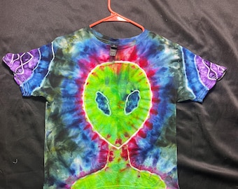 Puede incluir: Una camiseta teñida con un diseño de alienígena verde sobre un fondo colorido. El alienígena tiene ojos grandes y una boca pequeña. Los colores de la camiseta incluyen verde, azul, morado y rojo. La camiseta está colgada en una percha roja.