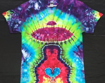 Puede incluir: Una camiseta teñida con un diseño vibrante que presenta una figura central con un corazón, rodeada por una forma similar a un OVNI. Los colores de la camiseta incluyen morado, verde, azul y rojo, creando un efecto psicodélico. La camiseta está colgada en una percha blanca.