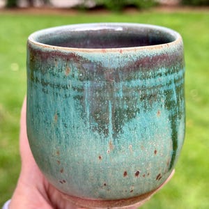 May include: A ceramic cup with a rounded shape and a glossy glaze in shades of teal, green, and brown. The surface features a mottled texture with dark brown speckles and a darker rim, showcasing a reactive finish with visible color variations.