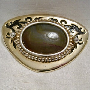 May include: A gold-toned metal belt buckle with a brown agate stone set in a silver-toned oval. The buckle is decorated with black floral designs.