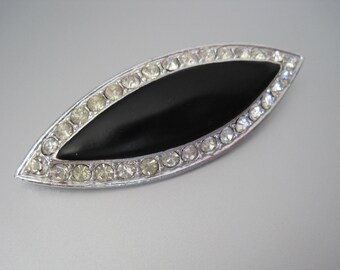 May include: A vintage brooch with a silver-tone metal frame. The brooch features a central black, oval-shaped insert, encircled by a row of clear, sparkling rhinestones. The design is symmetrical and elegant, suitable for formal occasions.