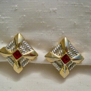 Könnte beinhalten: Ein Paar gold- und silberfarbene quadratische Ohrringe mit einem roten quadratischen Edelstein in der Mitte. Die Ohrringe haben ein strukturiertes, gewebtes Design und sind vor einem weißen Hintergrund platziert.