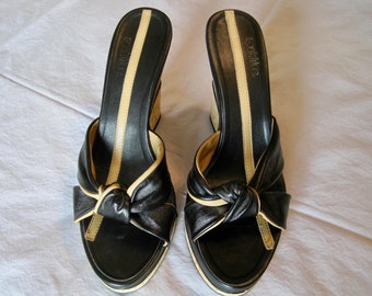 May include: A pair of black leather wedge sandals with a beige trim and a bow detail on the front. The sandals have the brand name "Sofft" written on the side.