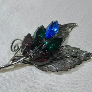 May include: A silver-toned leaf brooch with textured detailing. The brooch features a cluster of colourful, faceted gemstones in shades of blue, green, and purple. A vintage accessory.
