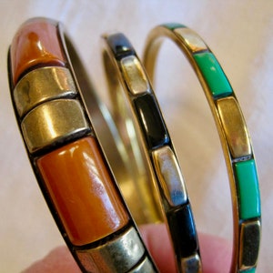 Peut inclure: Trois bracelets de différentes couleurs. L'un présente des segments rectangulaires orange avec des accents dorés et argentés. Un autre a des segments noirs, et le troisième a des segments verts. Tous sont sertis dans du métal doré.