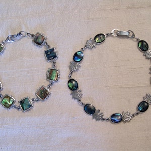 May include: Two silver bracelets with iridescent abalone shell accents. One bracelet has square shaped shells and the other has oval shaped shells.