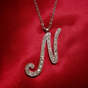 May include: A silver necklace with a large, cursive letter N pendant encrusted with clear rhinestones. The necklace is displayed on a red satin background.