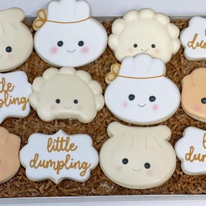 May include: A dozen decorated sugar cookies in a box. The cookies are shaped like dumplings with smiling faces and blush. Some cookies are white, some are tan, and some have the words "little dumpling" in gold script.