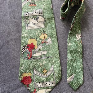 Peut inclure: Cravate verte avec un motif répété de notes de musique et de personnages de Peanuts. La cravate a un fond vert clair avec des notes de musique blanches et des illustrations de Snoopy, Charlie Brown et d'autres personnages.