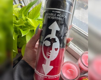 May include: A glass candle with a black top and red base. The candle features the words "ALLEGED DOUBLE ACTION REVERSIBLE" and a white graphic of a face and arrow. Made by Trader Candle Co., Brooklyn, NY.
