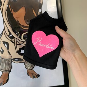 Puede incluir: Camiseta negra para perro con un coraz&oacute;n rosa de purpurina que dice "Barbie".