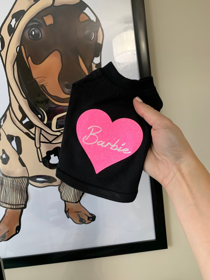 Puede incluir: Camiseta negra para perro con un coraz&oacute;n rosa de purpurina que dice "Barbie".
