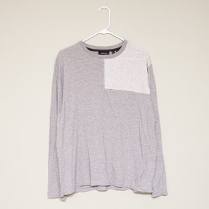 May include: A long-sleeved grey t-shirt with a white panel on the right shoulder. The shirt has a crew neck and is made of a soft, comfortable fabric.