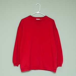 May include: A solid red sweatshirt with a crew neck and long sleeves. The sweatshirt is hanging on a white hanger against a light green wall.