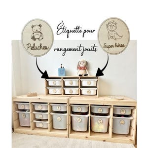 Peut inclure: Meuble de rangement de jouets en bois avec plusieurs bacs blancs. Les bacs sont étiquetés avec des étiquettes en bois. Les étiquettes incluent les mots "Peluche" et "Super héros". Le meuble est en bois clair et est conçu pour ranger les jouets.