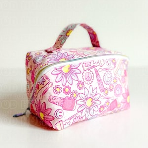 May include: A pink cosmetic bag with a white zipper and a matching handle. The bag is covered in a pattern of pink flowers, peace signs, lipstick, and nail polish bottles. The bag is designed to hold makeup and other beauty products.