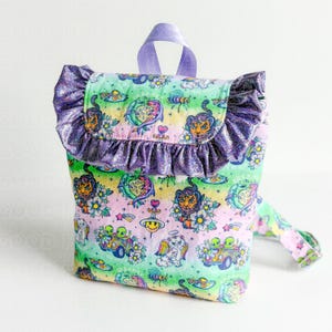 May include: A backpack featuring a pastel rainbow gradient background with a repeating pattern of tigers, aliens in cars, cats with wings, and flowers. The bag has a purple glittery ruffled trim along the top flap and a purple fabric handle.