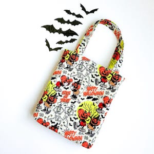 Pu&ograve; includere: Una borsa in tessuto con un motivo ripetuto di Halloween composto da personaggi con testa di zucca arancione, scheletri bianchi, pipistrelli neri e la scritta "Happy Halloween" e "Trick or Treat" in arancione su sfondo bianco.