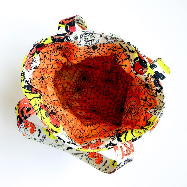 Pu&ograve; includere: Una borsa in tessuto con una stampa a tema Halloween raffigurante scheletri, zucche e pipistrelli su sfondo bianco. L'interno &egrave; foderato con un tessuto arancione brillante decorato con un motivo a ragnatela nera e due manici robusti.
