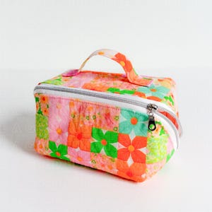 May include: A rectangular fabric cosmetic bag featuring a quilted patchwork pattern of orange, pink, and green flowers. The bag includes a top handle, a white zipper closure with a silver metal pull, and a structured shape for storage.