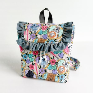 May include: A small backpack with a colorful Star Wars-themed pattern. The design includes images of R2-D2, BB-8, Chewbacca, and other characters, along with flowers and hearts. The backpack has a gray ruffle trim and a drawstring closure.