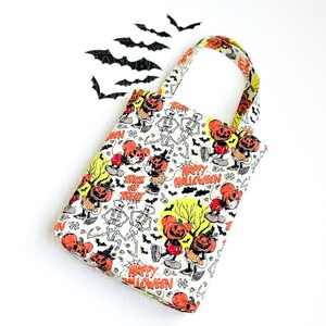 Pu&ograve; includere: Una borsa in tessuto bianco con un motivo ripetuto di Halloween che presenta personaggi con teste di zucca arancioni, scheletri danzanti, pipistrelli neri e alberi gialli. Il testo Happy Halloween e Trick or Treat &egrave; stampato in arancione.
