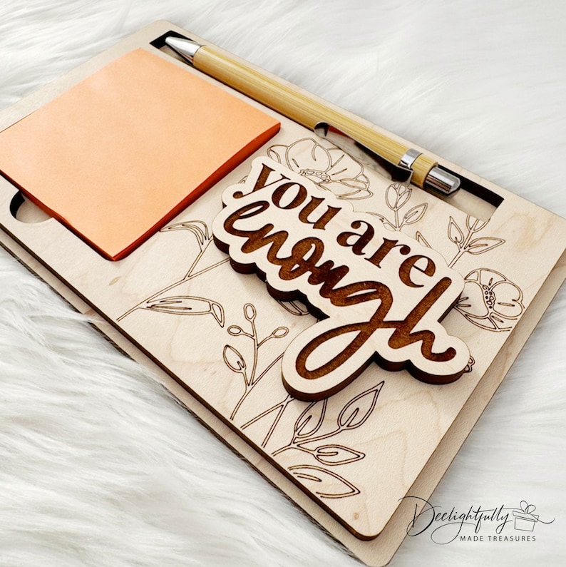 May include: Wooden desk organizer with a floral design and the words "You are enough." Includes a notepad and a bamboo pen. The organizer is light brown with etched floral details and a light orange notepad.