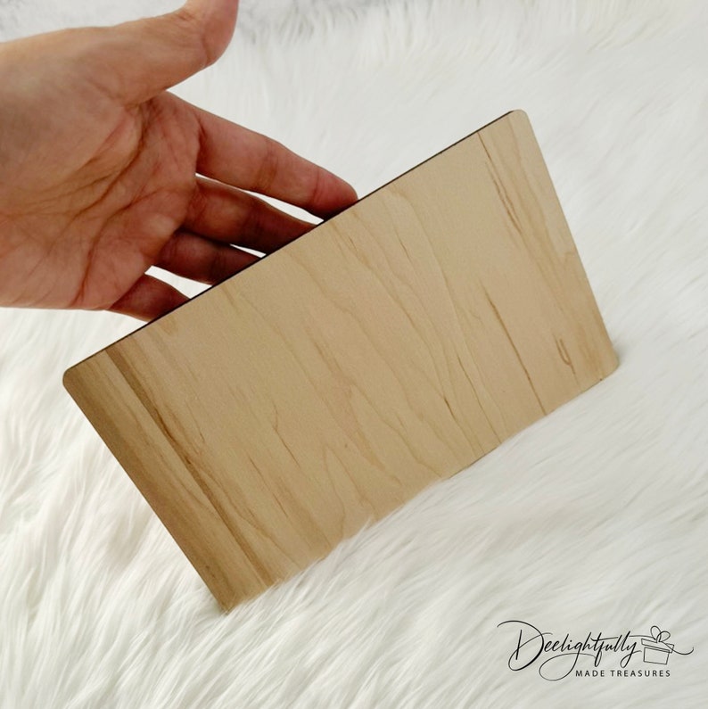 May include: A rectangular wooden plaque with a light brown finish. The plaque has rounded corners and a dark edge. The surface shows the natural grain of the wood. The plaque is held by a hand.