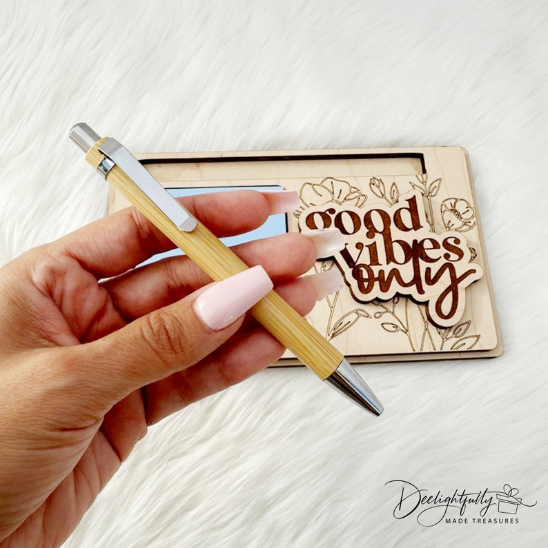 May include: A bamboo pen with silver accents is held in front of a wooden sign. The sign has the words "good vibes only" in a brown font, with floral designs. The pen is light brown and silver.