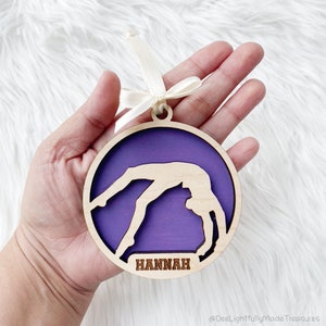 May include: A round wooden ornament with a gymnast silhouette in a bridge pose, set against a purple background. The ornament has a natural wood frame and the name "HANNAH" is engraved at the bottom. A cream-colored ribbon is attached for hanging.