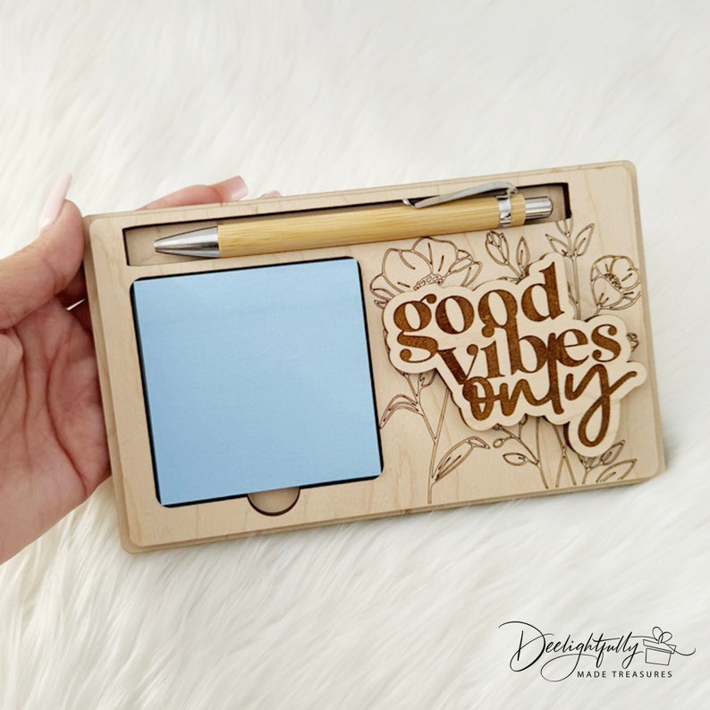 May include: Wooden desk organizer with a light blue sticky note pad, a bamboo pen, and the phrase "good vibes only" in a decorative font. The organizer has floral engravings.