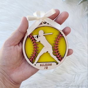 May include: A wooden softball ornament with a yellow center and pink stitching detail. A silhouette of a batter is cut out of the wood. The name "Allison" and the number "#8" are engraved on the ornament. A cream-colored ribbon is attached for hanging.