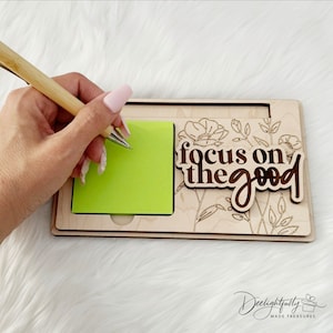 May include: A wooden desk organizer with a green sticky note pad and a bamboo pen. The organizer has a floral design and the words "focus on the good". The pen is held by a person.