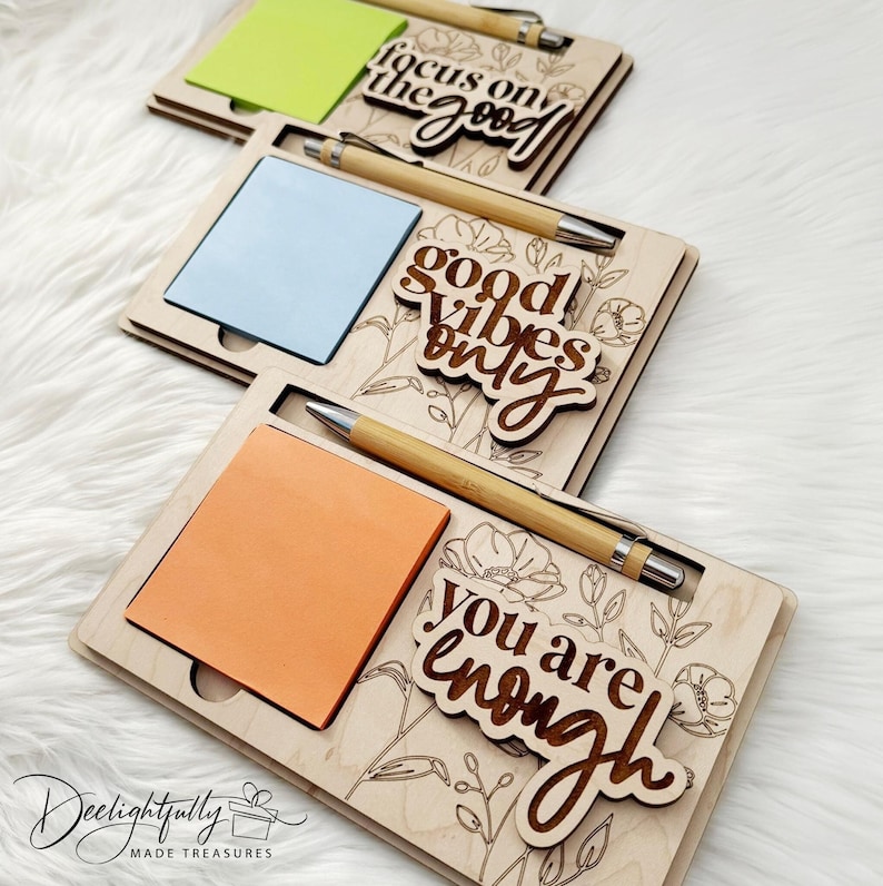 May include: Three wooden memo pads with pens, each holding a different colored sticky note. The pads have floral designs and phrases like "focus on the good," "good vibes only," and "you are enough." The pens have bamboo barrels.