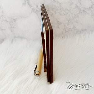 May include: A wooden sign with a dark brown frame and a light blue interior. A light-colored pen rests against the sign. The sign is on a white, fluffy surface. The sign has the text "Deelightfully Made Treasures" in cursive.