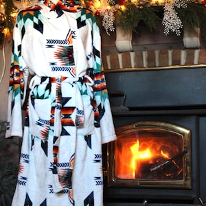 May include: A white hooded robe with a colorful geometric pattern in shades of blue, orange, green, and black. The robe has a matching belt and pockets. The robe is displayed on a mannequin in front of a fireplace.