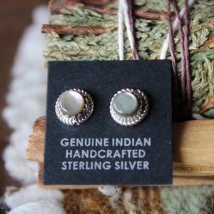 Pode incluir: Dois brincos redondos com centros de madrepérola iridescente em molduras de prata esterlina trançada. Os brincos estão fixados num cartão preto impresso com o texto GENUINE INDIAN HANDCRAFTED STERLING SILVER.