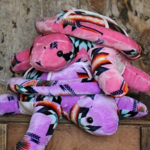 May include: A stack of plush turtle toys in shades of pink and purple. The turtles have black eyes and are covered in a patterned fabric with geometric designs in orange, black, and white.