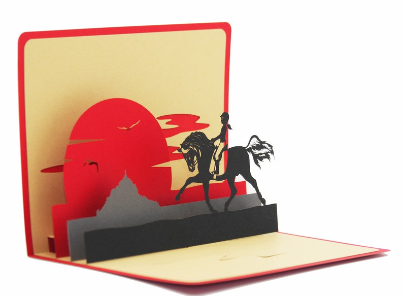 May include: A red, black, and gray 3D pop-up card with a silhouette of a person riding a horse in front of a red sun and gray mountains.