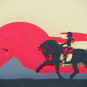 May include: A black silhouette of a person riding a horse against a red sunset background. The horse is rearing up on its hind legs. The person is wearing a helmet and riding in a riding position. The background is a red sunset with a gray mountain in the foreground.