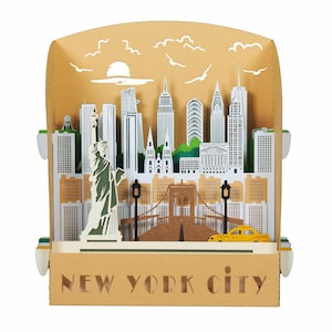 Peut inclure: Diorama en papier 3D de New York City, avec des monuments emblématiques comme la Statue de la Liberté, l'Empire State Building et le pont de Brooklyn. Le diorama est dans une boîte beige avec les mots "NEW YORK CITY" en bas. Inclut un taxi jaune.